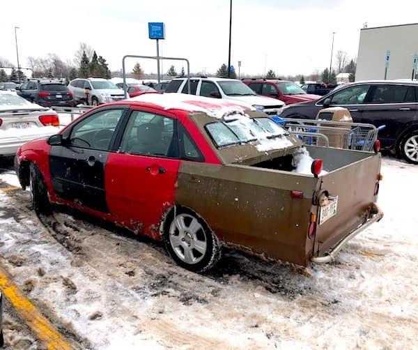 A winter-ready redneck DIY car spotted in a snowy parking lot. A red four-door sedan has had its trunk removed and replaced with a custom-built, brown metal pickup bed, complete with its own taillights and a rear-facing window.