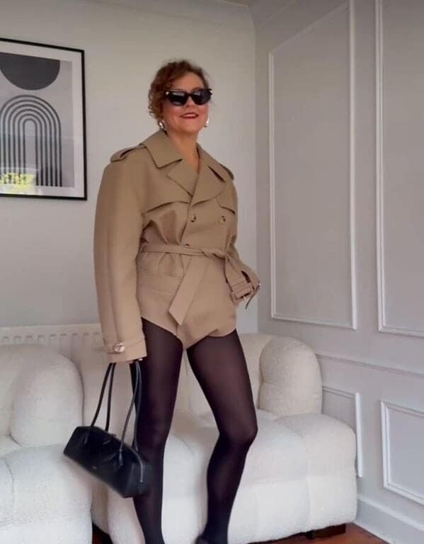street-style photo of a woman wearing a "no pants" look consisting of black opaque tights and a tan double-breasted trench coat that has been cropped so high it functions as a bodysuit.