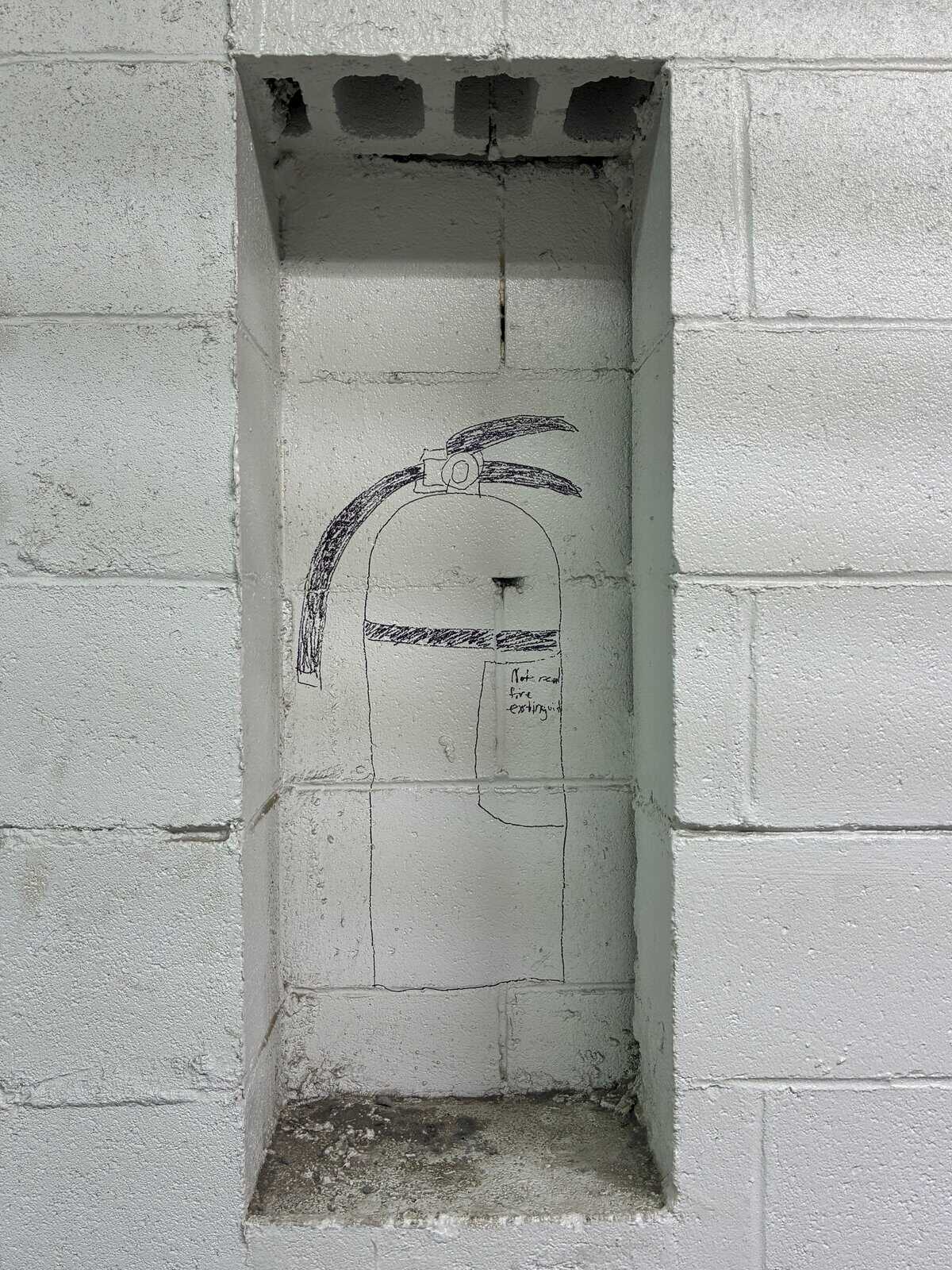 minimalist act of wholesome vandalism inside a recessed wall niche. Instead of a real safety device, a crude, hand-drawn sketch of a fire extinguisher occupies the space, accompanied by the small, honest note: "Not a real fire extinguisher."