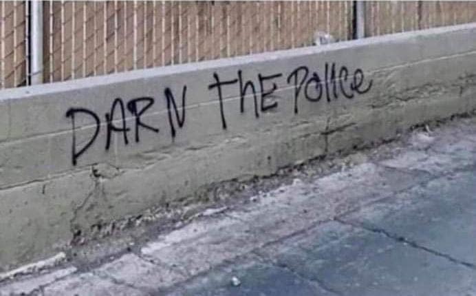 concrete wall featuring a remarkably polite act of rebellion. Spray-painted in black capital letters is the phrase "DARN THE POLICE," offering a G-rated, extremely mild alternative to a more common and aggressive anti-authority slogan.