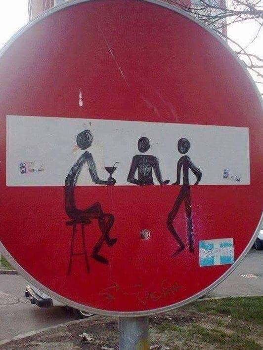 A circular red "No Entry" street sign featuring a white horizontal bar. Creative wholesome vandalism has transformed the bar into a countertop, with black marker stick figures sitting on stools and standing around, including one holding a tiny martini glass.