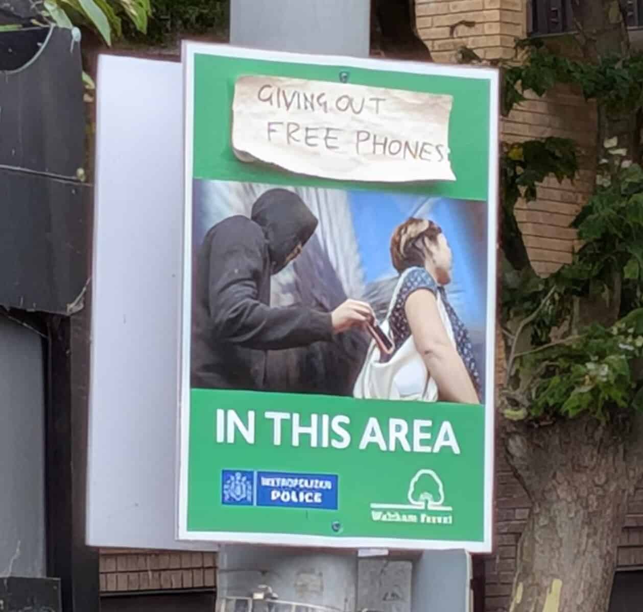 A Metropolitan Police poster warning about pickpockets, showing a hooded man reaching into a woman's bag to take her phone. Wholesome vandalism has added a handwritten note at the top that recontextualizes the crime as a charity: "GIVING OUT FREE PHONES."