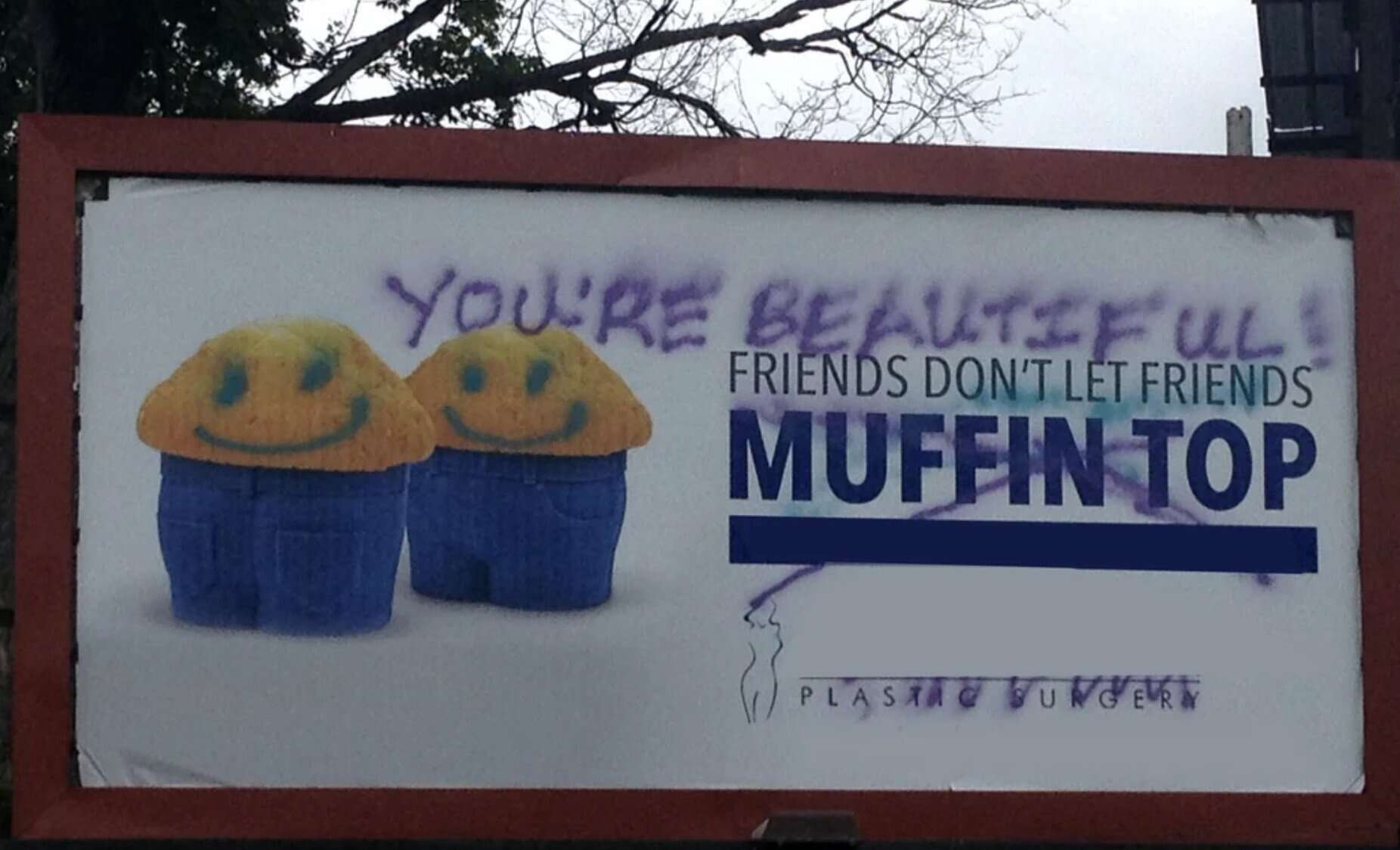 billboard for plastic surgery featuring muffins wearing blue jeans to illustrate a "MUFFIN TOP." Vandalized with a positive message, purple spray paint reads "YOU'RE BEAUTIFUL!" across the top, and blue smiley faces have been added to the muffins
