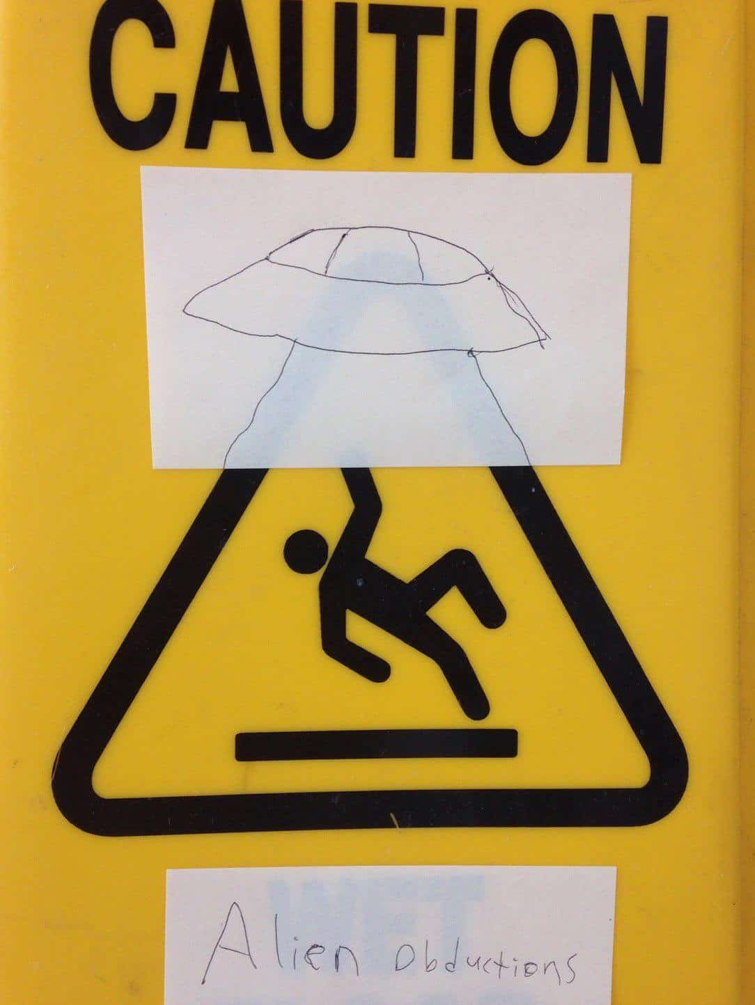 A yellow "Caution" sign for a wet floor showing a falling stick figure. Clever wholesome vandalism uses a paper cutout of a UFO positioned above the figure, turning a standard slip-and-fall warning into an "Alien abductions" alert.