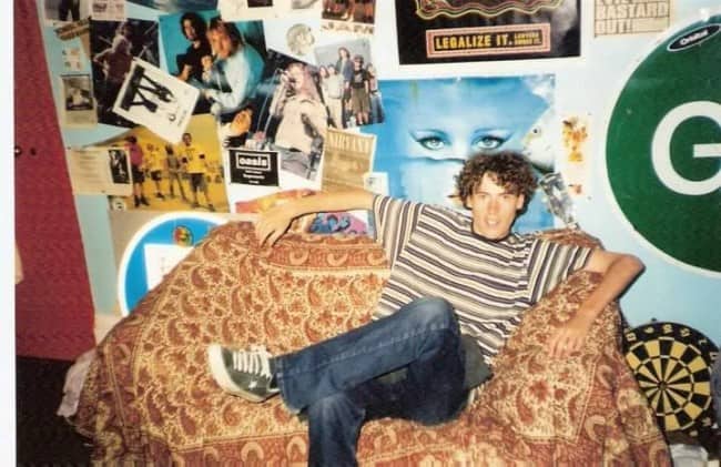 Curly haired teen lounging in a papasan chair in front of a wall of 90s posters.