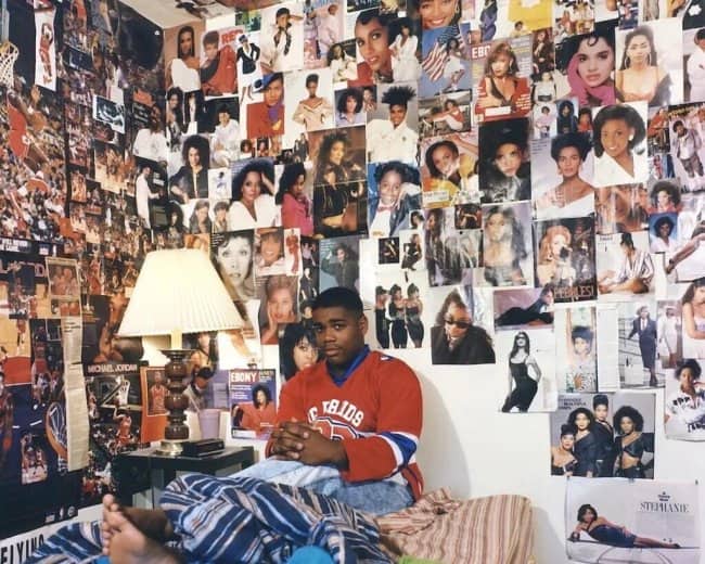 Teen in a red jersey sitting on his bed beneath walls covered in magazine photos.