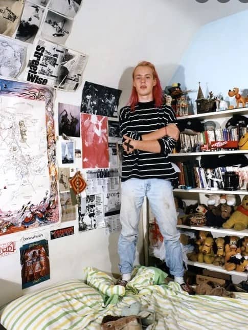 Teen with pink hair standing on a bed next to a wall of zines and plushies.