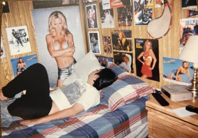 Teen lying on a bed under walls covered in pinup and sports posters.