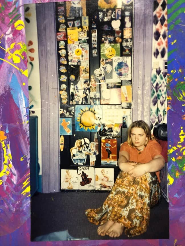 Teen in a floral skirt sitting before a door plastered in stickers and clippings.