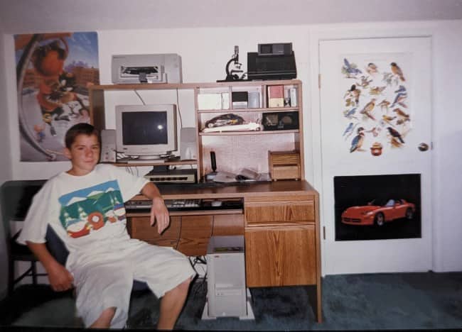 Boy in a white tee at a computer desk beside a microscope and Corvette poster.