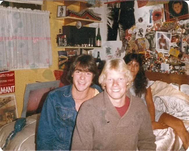 Three friends grinning on a bed in a cluttered bedroom covered in pins and posters.