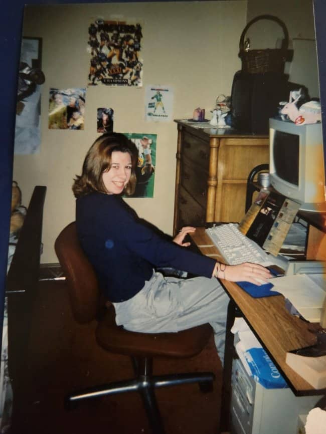 Smiling young woman at a desktop computer in a 90s bedroom with football posters.