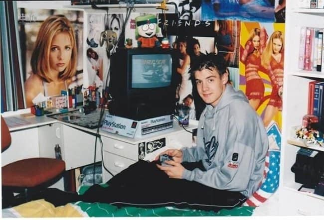 Teen in a gray hoodie playing PlayStation on his bed, surrounded by TV show posters.