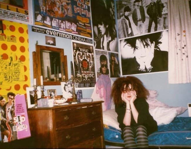 Teen with big curly hair on a bed surrounded by punk and goth band posters.