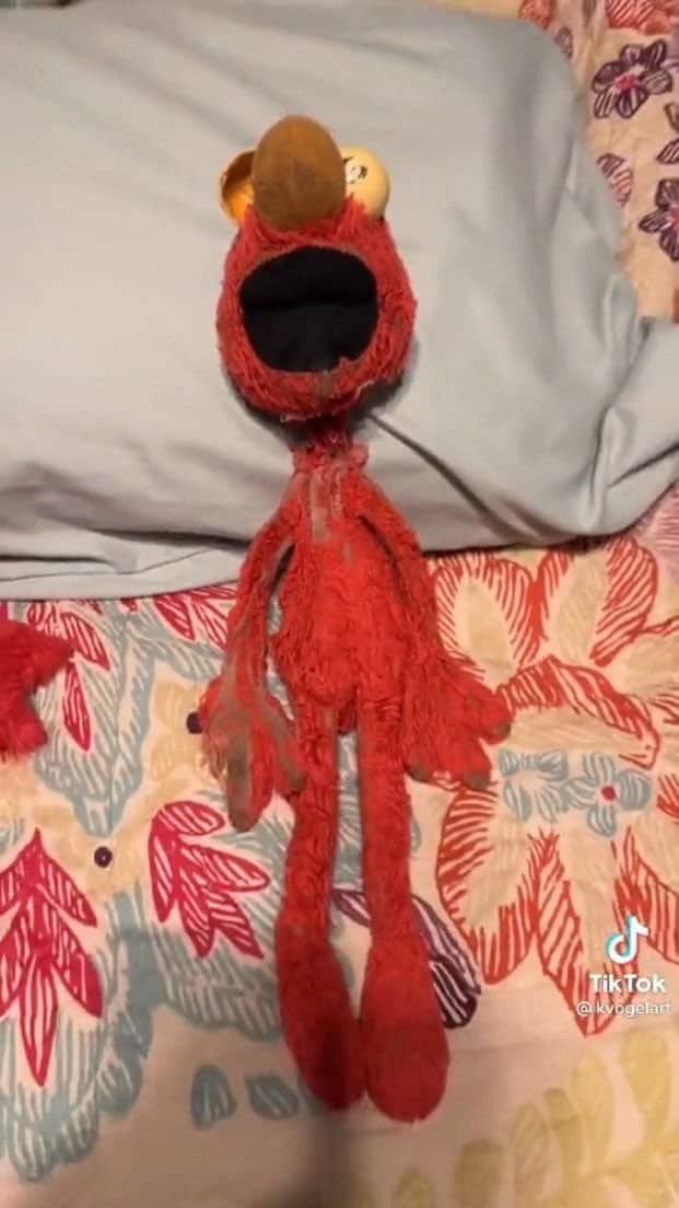 Worn out, stretched Elmo plush with a gaping mouth lying flat on a floral bedspread
