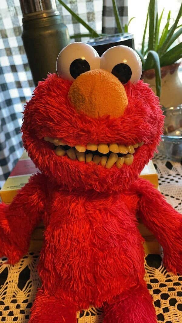 Elmo plush with a grin of realistic yellow human teeth sitting on a lace tablecloth