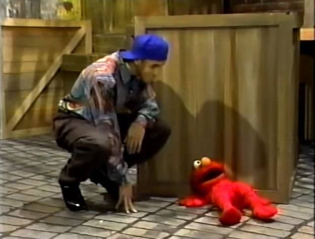 Man in a blue cap crouching beside an Elmo puppet lying face down on set