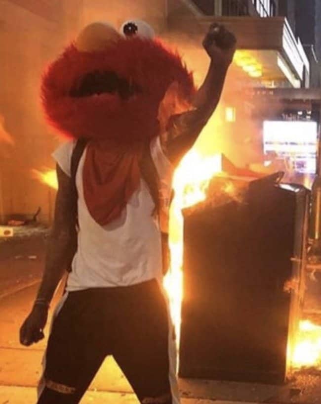 Person in an Elmo mask raising a fist in front of a burning dumpster at night
