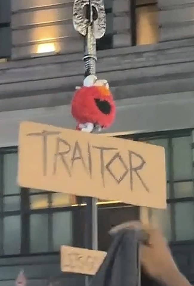 Small Elmo plush hanging from a pole above a handwritten TRAITOR sign at a street protest
