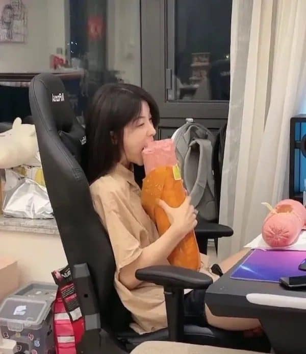 Woman in a gaming chair biting into an enormous orange sausage-shaped snack.