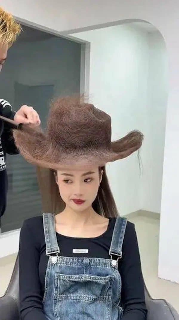A stylist combs a woman's hair into the shape of a giant brown cowboy hat.