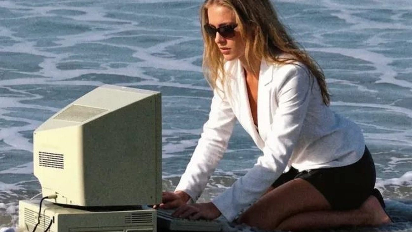 Woman in a white blazer typing on a beige desktop computer set up in ocean surf, a morning memes favorite about working remotely